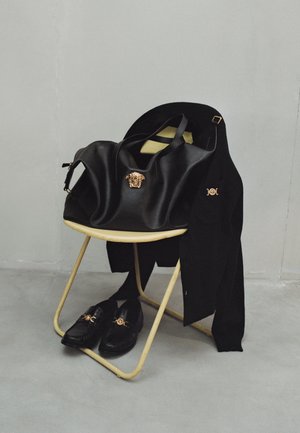 Zwarte leren schoudertas met gouden leeuwembleem, gecombineerd met zwarte loafers met gouden accenten, rustend op een lichtgele stoel. Zwarte trui over de stoel gedrapeerd.