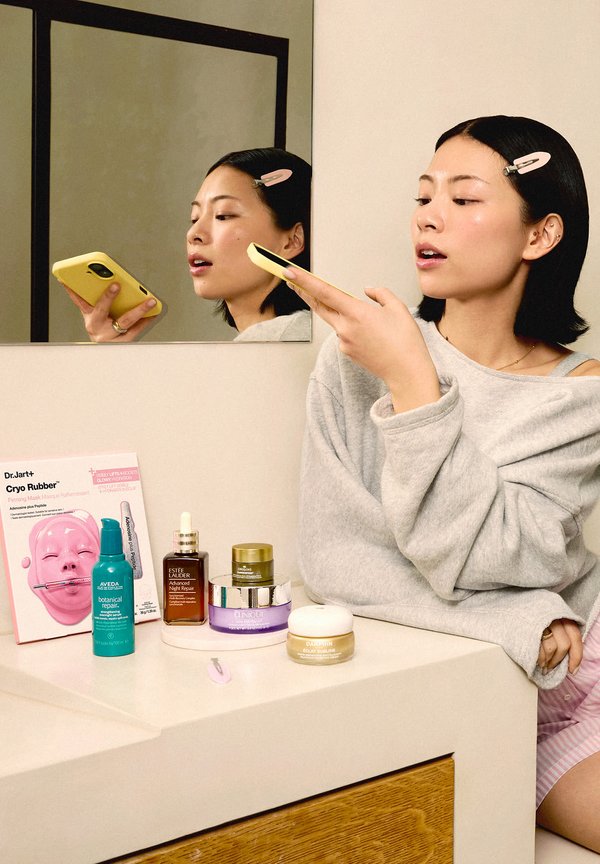 Vrouw met haarspeld die in een gele smartphone spreekt, zittend bij een badkamermeubel met verschillende huidverzorgingsproducten erop uitgestald.