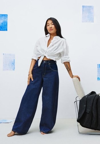Model in een wit geknoopt shirt en donkerblauwe wijde denim jeans. Op de achtergrond is lichte blauwe kunst te zien op een witte muur.