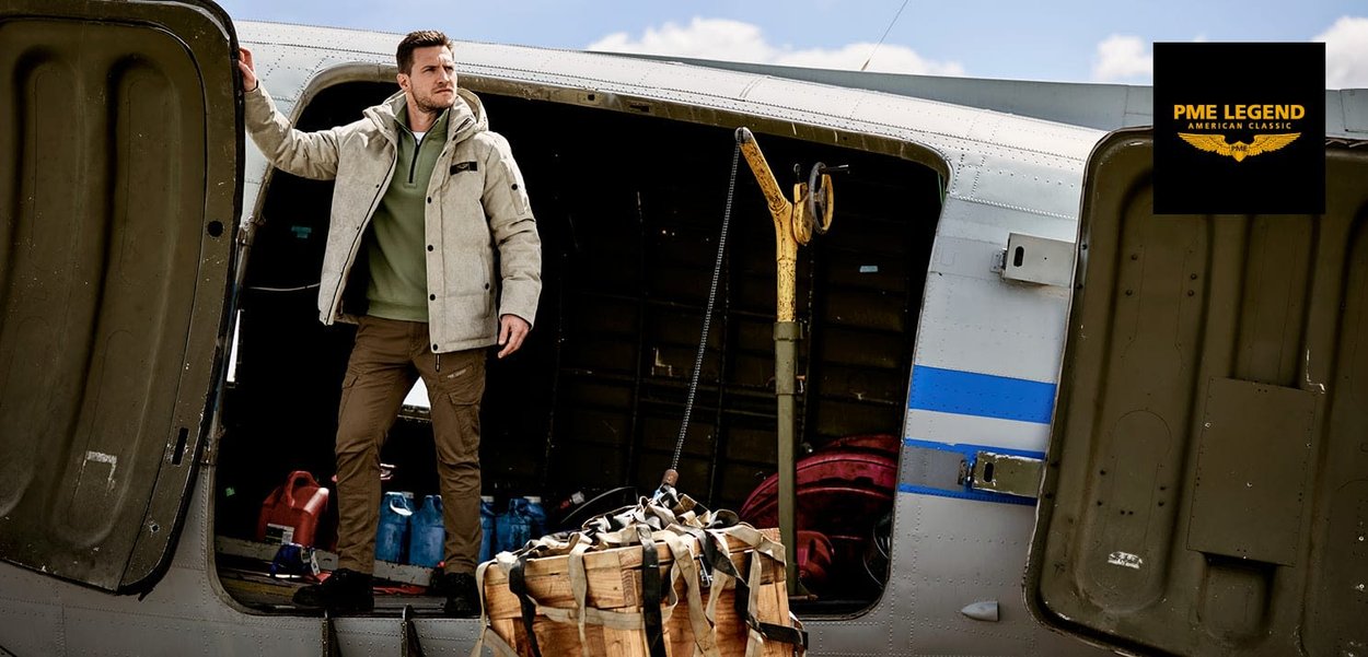 Veste isolante grise sur un pull vert, pantalon cargo brun, debout dans une porte d'avion ouverte. Une caisse en bois est suspendue en dessous.