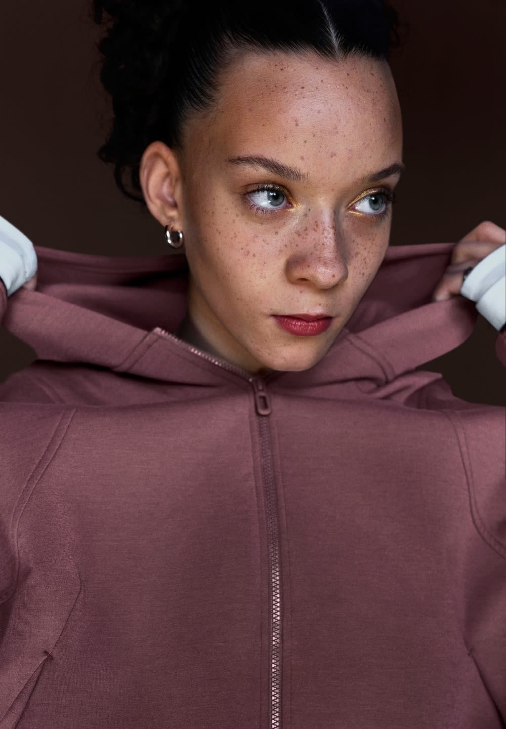Person med fregner og hoops-øreringe justerer hætten på en lavendelfarvet lynlåssweatshirt mod en mørk baggrund.