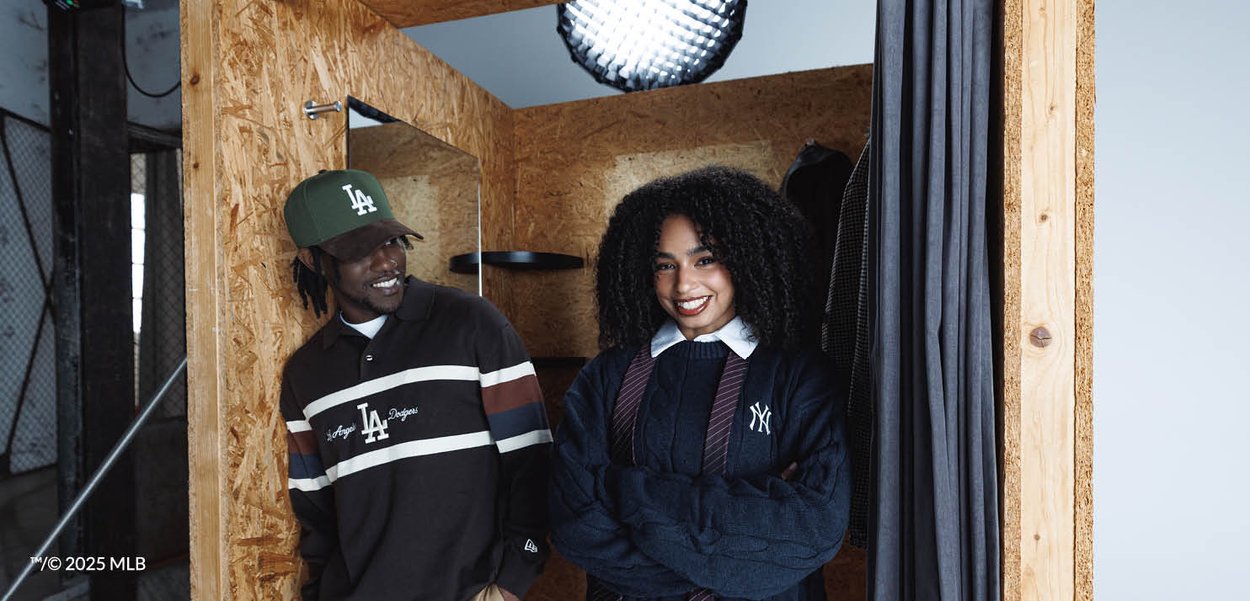 To personer smilende i en prøverum af træ, den ene iført en Dodgers-kasket og sweater, den anden iført en Yankees-sweater med seler.