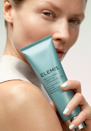 Kvinne med blekeblå neglelakk holder en blå tube med Elemis Pro-Collagen Hydrating Cleansing Mousse nær skulderen, vendt mot kameraet.