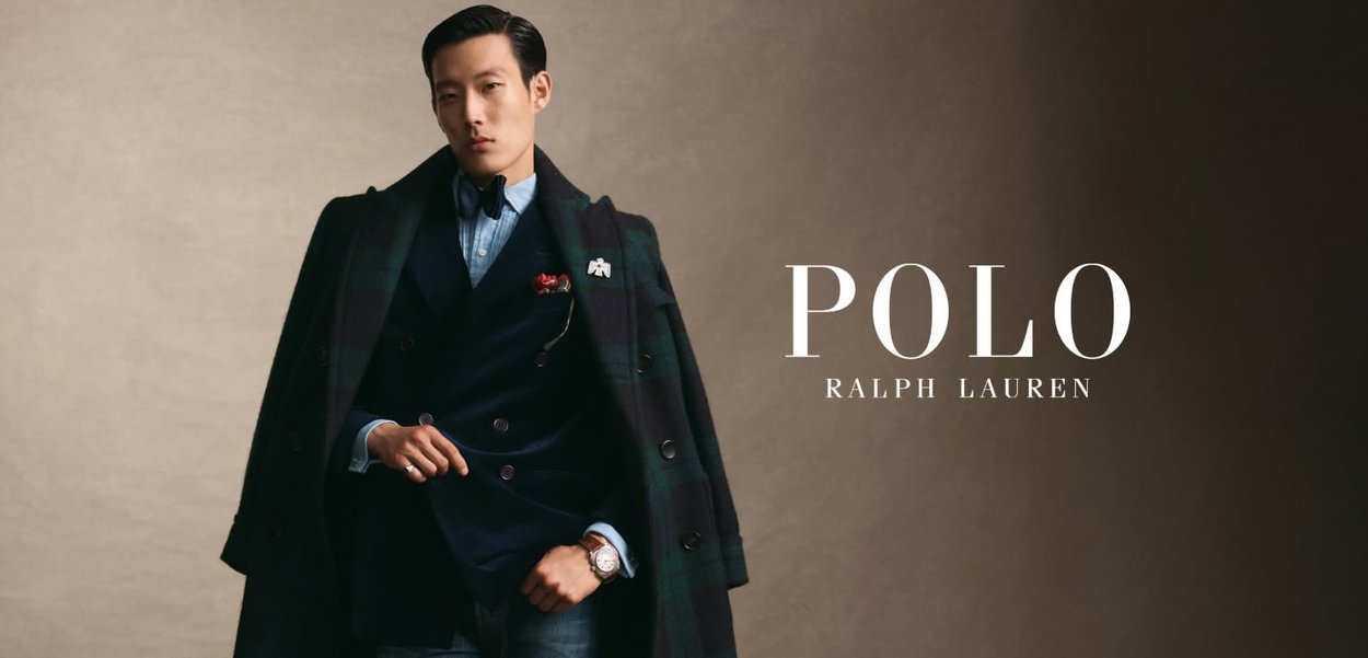 Manteau à carreaux long sur un blazer bleu marine, une chemise bleue et un jean foncé. Les accessoires incluent une broche et une montre. L'arrière-plan est neutre et texturé.