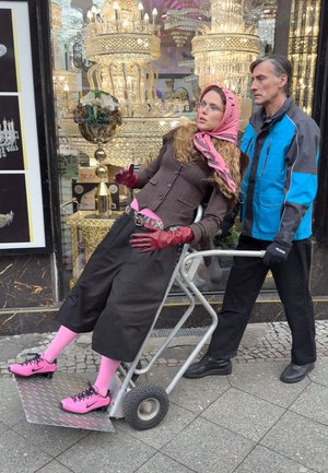 Zwei Personen in der Nähe eines Ladengeschäfts. Eine lehnt an einem Metallwagen, trägt einen rosa Schal, einen langen braunen Mantel und rosa Socken mit schwarzen Turnschuhen.
