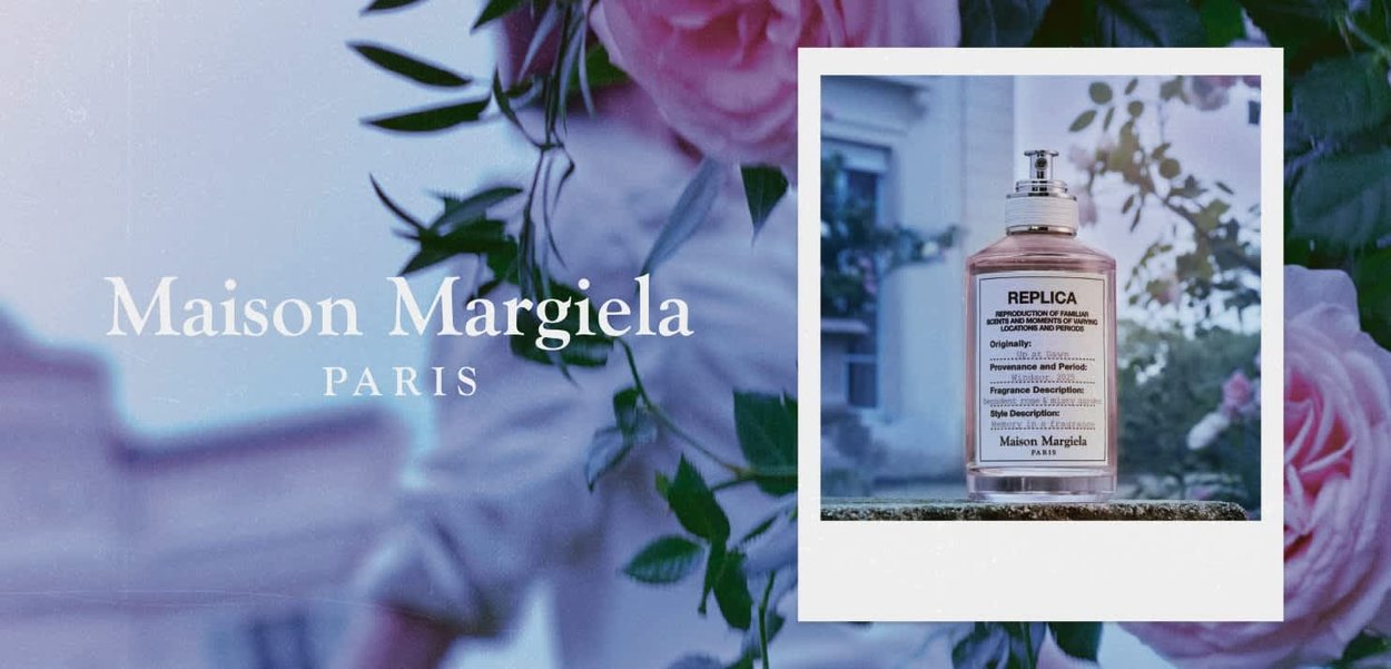 Bottiglia del profumo Maison Margiela Replica su una superficie con rose sfocate e edificio sullo sfondo.