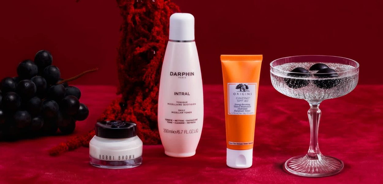 Drie cosmetische producten: een witte fles met het etiket "Darphin Intral," een oranje tube "Origins Ginzing SPF 40," en een klein zwart potje. Een glas met zwarte druiven en bubbels.