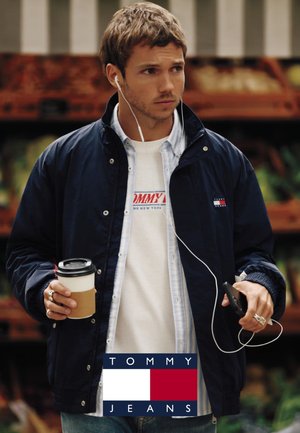 Giovane uomo indossa una giacca Tommy Jeans blu navy e una maglietta bianca, tiene in mano una tazza di caffè e uno smartphone con cuffie in un ambiente all'aperto.