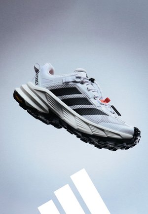 Zapatilla blanca de trail running Adidas Terrex con rayas negras y suela de taco negra gruesa flotando sobre un fondo gris claro.