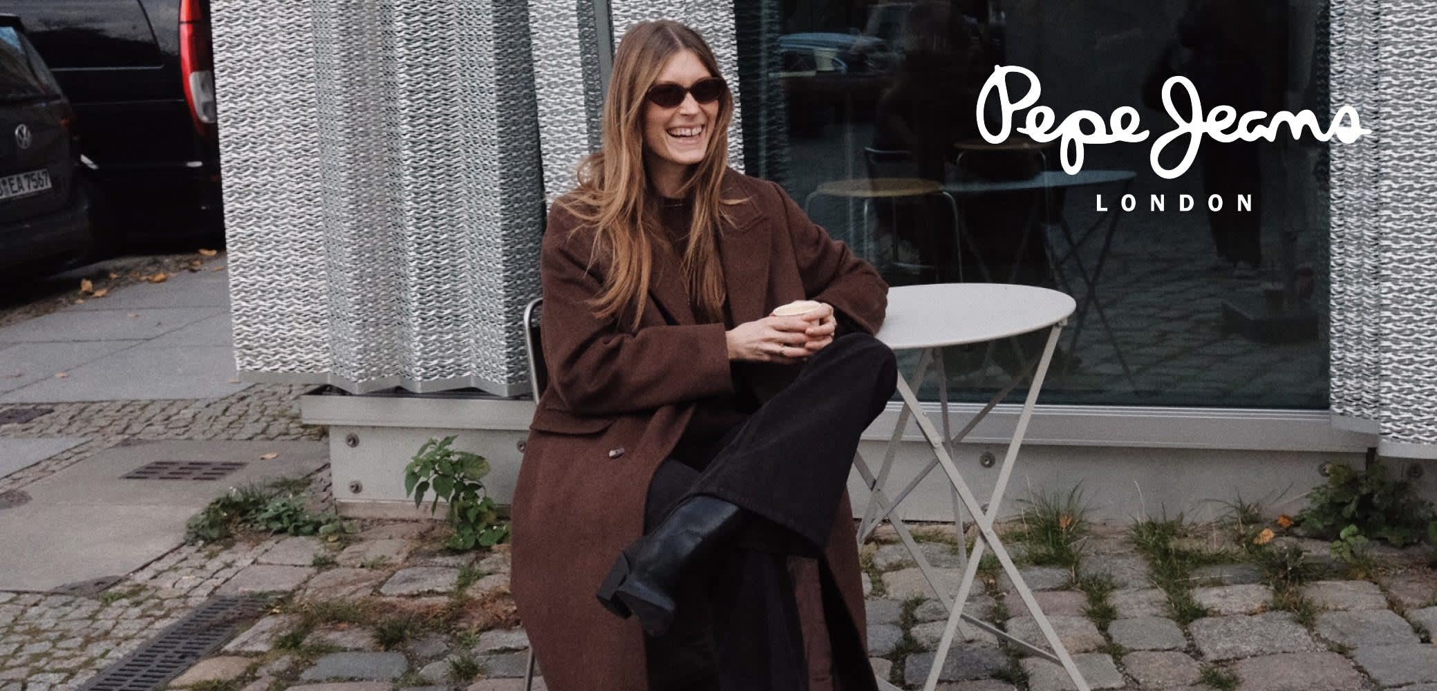 Abrigo marrón de gran tamaño, pantalones negros de pierna ancha y botas negras hasta la rodilla. Modelo sentada en una mesa blanca pequeña junto a una pared plateada con textura.