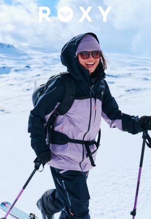 Lila-schwarze wasserdichte Skijacke mit Reißverschlüssen, Taschen-Details und verstellbaren Riemen. Getragen mit Skistöcken auf schneebedecktem Gelände.