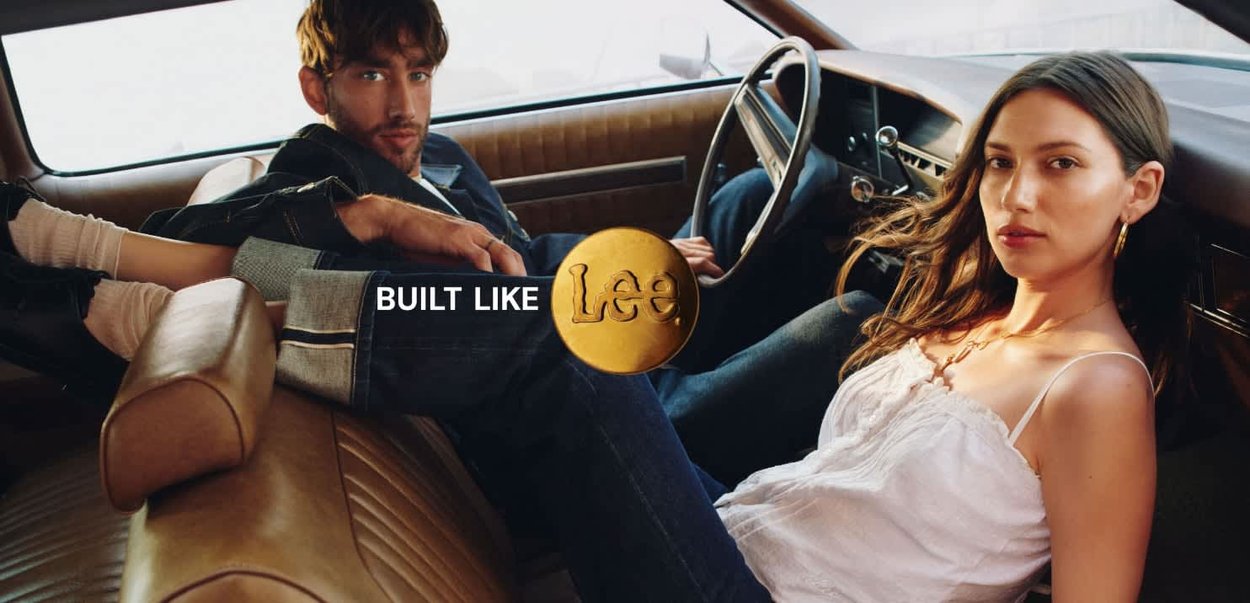 Et vintage bilinteriør har brunt lærpolstring. To personer sitter på plassene, med et synlig gull-emblem "Built Like Lee" i midten.