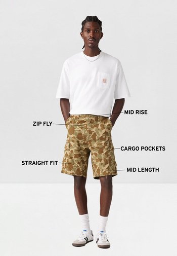Shorts cargo camuflados con cintura de altura media, corte recto, cremalleras, longitud media y múltiples bolsillos. Combinados con una camisa blanca de manga corta.