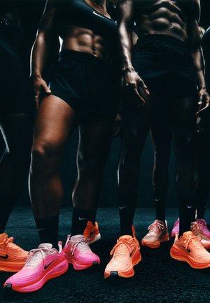 Nike Sportschuhe in Pink- und Orangetönen zeichnen sich durch ein schlankes Design, eine strukturierte Oberfläche und logoplatzierungen aus und werden von Personen in schwarzer Kleidung getragen.