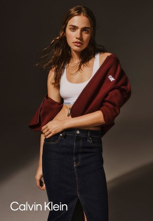 Burgundowa bluza z zamkiem błyskawicznym z podwiniętymi rękawami, biała crop top i ciemnoniebieska spódnica dżinsowa z rozcięciem z przodu. Logo Calvin Klein na wykończeniu.