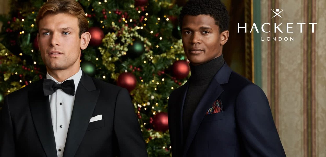 Twee mannen in formele kleding staan voor een versierde kerstachtergrond. De ene draagt een zwart smoking, de andere een donkere jas met een gestreepte pochet.