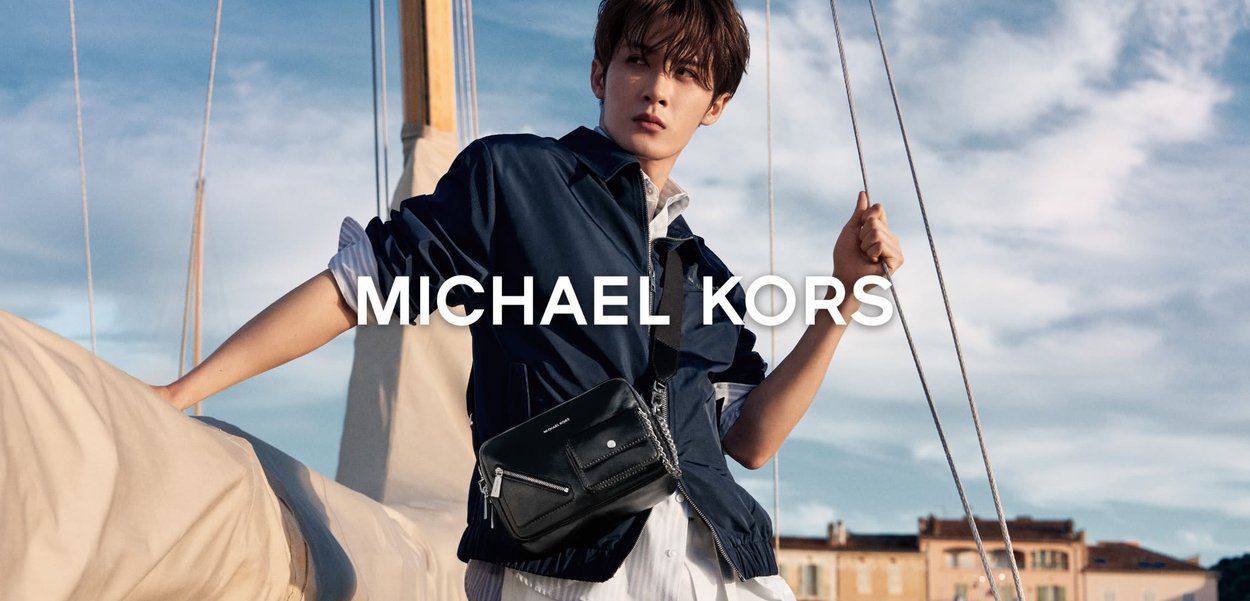 Jonge man in een marineblauw jasje en wit overhemd poseert terwijl hij zeilboottouwen vasthoudt, met een zwarte Michael Kors crossbodytas onder een blauwe lucht.