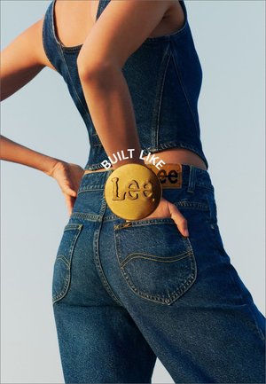 Mørkeblå denim jeans med en tætsiddende pasform og gule syninger. Har en stor, rund guld-bæltespænde med "Lee" præget.