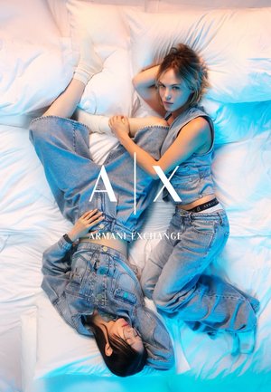 Dos mujeres con conjuntos de mezclilla acostadas sobre ropa de cama blanca, una reclinada con los ojos cerrados, la otra mirando a la cámara, con el logotipo de A|X Armani Exchange superpuesto.