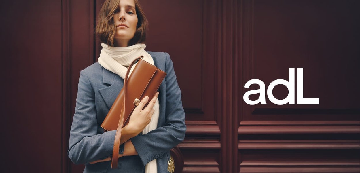 Lichte bruine leren handtas met een slank ontwerp, voorzien van een sluiting, vastgehouden door een persoon in een blauwe blazer en een crèmekleurige sjaal, tegen een maroen achtergrond.