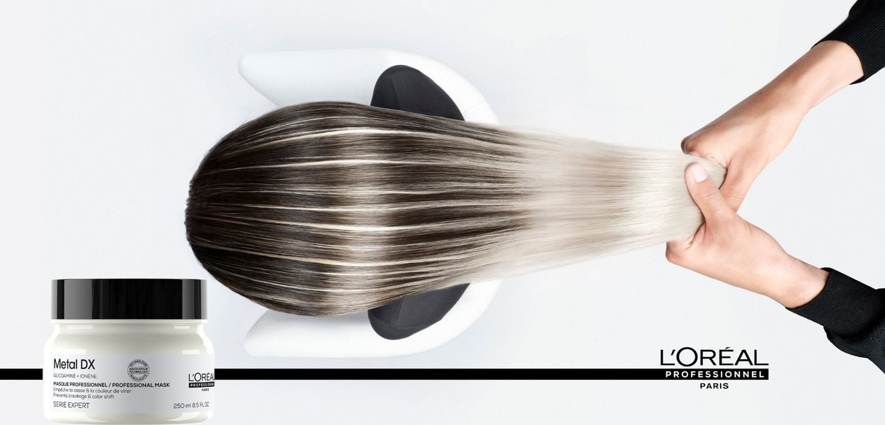 Mani che tengono capelli lunghi, lisci, sfumati da scuro a chiaro sopra un contenitore della maschera per capelli L'Oréal Professionnel Metal DX su uno sfondo bianco.