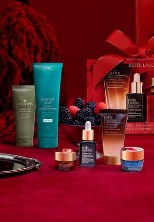 Prodotti per la cura della pelle su una superficie rossa: tubetto verde di Origins, tubetto blu di Aveda e diverse bottiglie di Estee Lauder con accenti dorati e una scatola regalo con un nastro rosso.