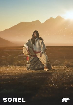 Sorel-Schuhe an einem Modell auf einem Felsen, das einen cremefarbenen Oversize-Pullover und helle Hosen trägt, vor einer Bergkulisse bei Sonnenuntergang.