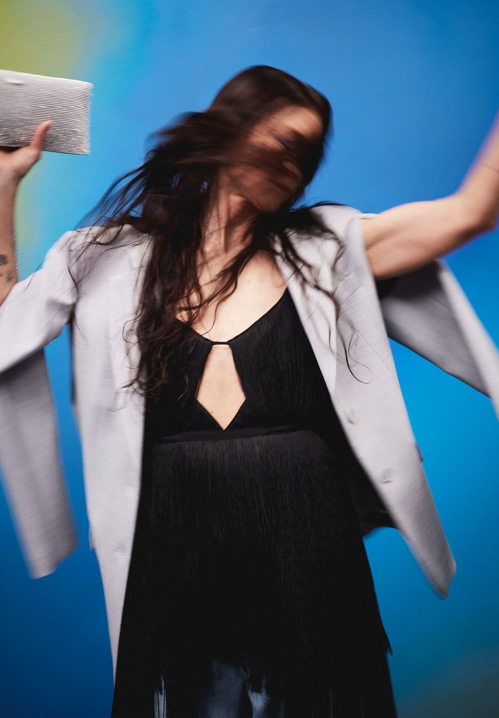 En flödande svart klänning med djup V-urringning, prydd med fransdetaljer. Tillsammans med en ljusgrå oversized blazer och en silverfärgad clutch. Blå bakgrund.