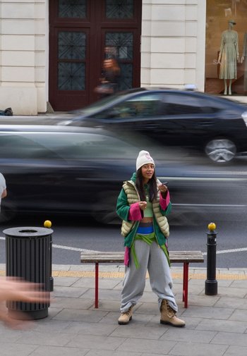 Jongere persoon in een groen en roze pufferjack over een lichtgroene top, grijze sweatpants en beige laarzen, staande op een stoep.
