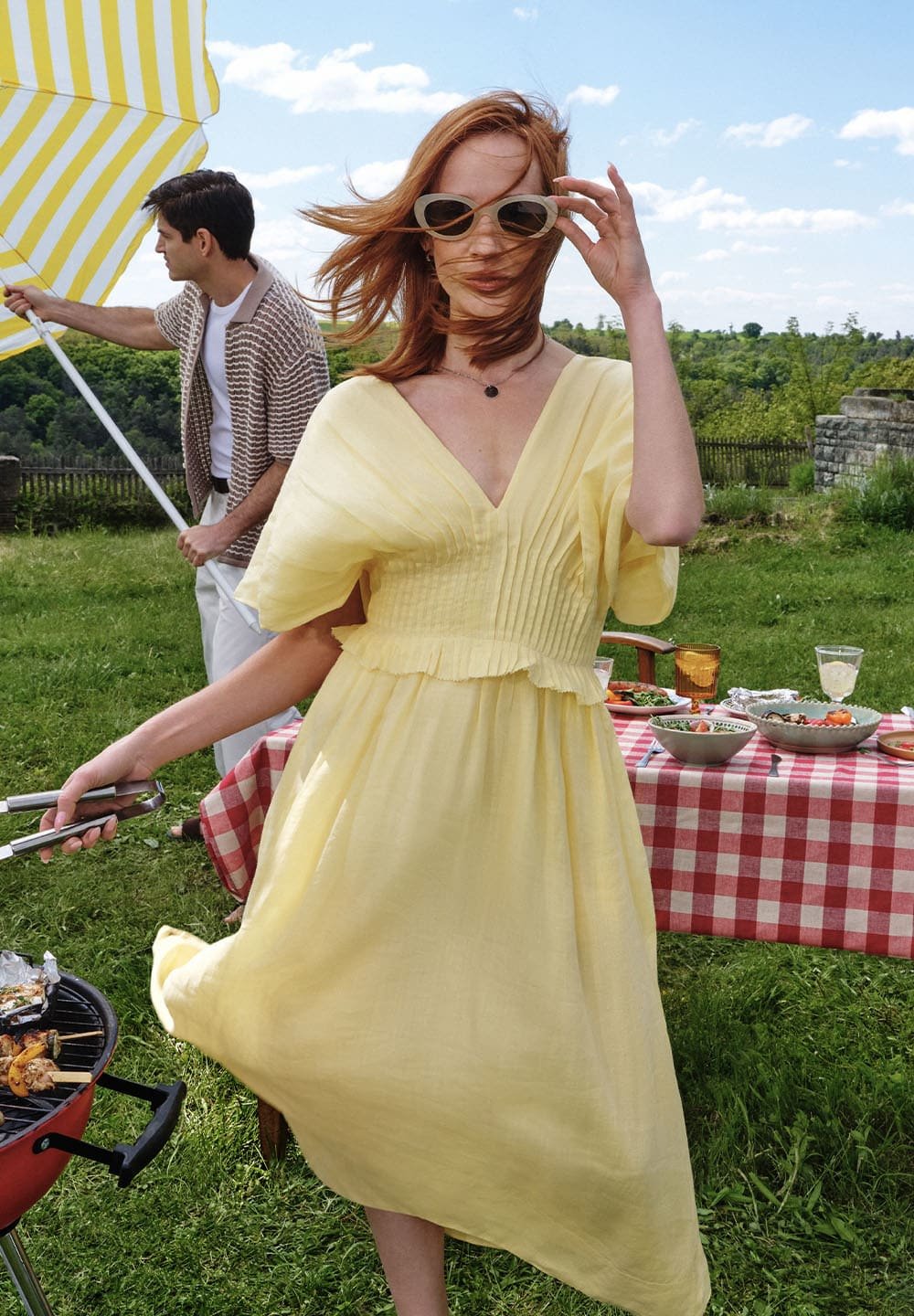 Žluté lněné šaty s krátkými nabíranými rukávy, výstřihem do V a skládaným korpusem. Modelka drží kleště, stojí na travnaté ploše s piknikovým uspořádáním.