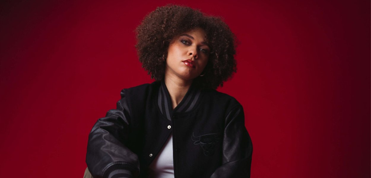 Czarna kurtka varsity z skórzanymi rękawami, żeberkowym kołnierzem, haftowanym logo na klatce piersiowej, noszona na białej bluzce na czerwonym tle.