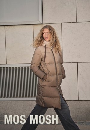 Long, beige puffer coat with a high collar and zipper closure. Features side pockets and vertical stitching. Model stands against a neutral background.