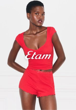 Vrouw staat met haar handen achter haar rug, gekleed in een rood T-shirt met korte mouwen en bijpassende rode short met kanten randje, met wit "Etam"-logo op de borst.