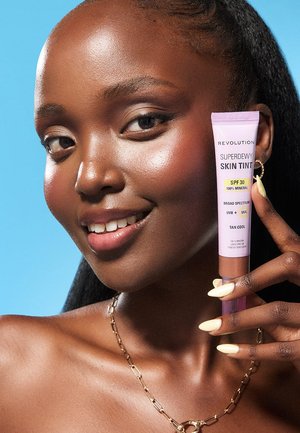 Donna sorridente con pelle luminosa che tiene un tubetto di tinta minerale per la pelle con SPF 30 su uno sfondo blu, mostrando trucco e unghie.