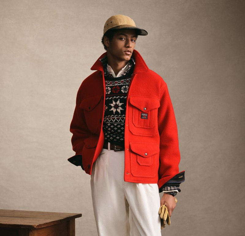 Jonge man met een rood jack, een gebreide trui met patroon, witte broek, beige pet en gele handschoenen in de hand, staand naast een houten tafel.