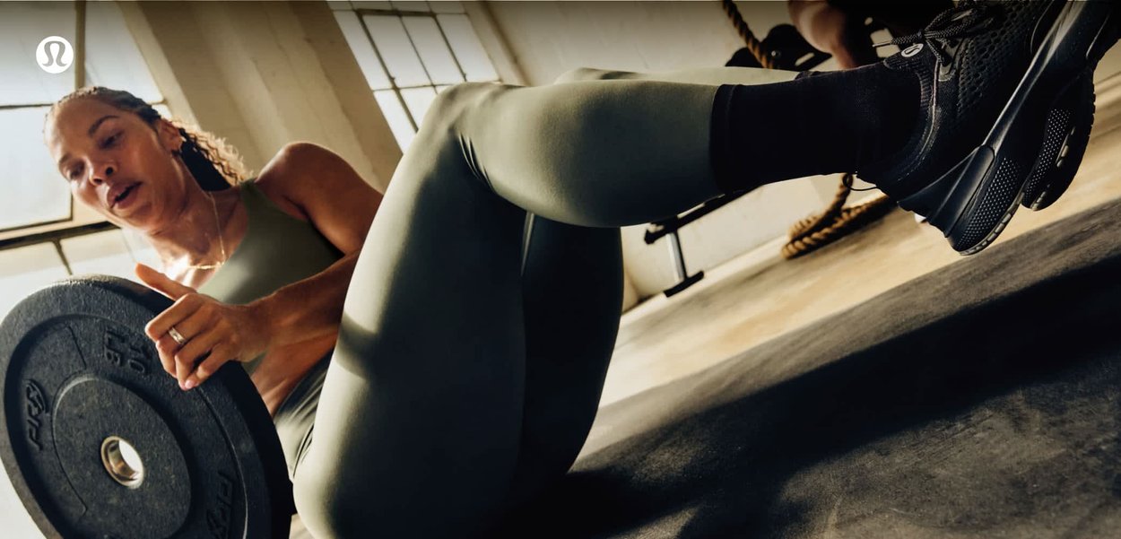 Frau in grüner Sportkleidung, die eine 10-Pfund-Hantelscheibe hält, sitzt mit angewinkelten Knien auf dem Boden in einem sonnenbeschienenen Fitnessstudio.