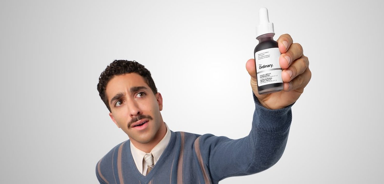Młody mężczyzna w swetrze trzyma i pokazuje butelkę z serum do pielęgnacji skóry oznaczoną "The Ordinary" na jednolitym jasnym tle.