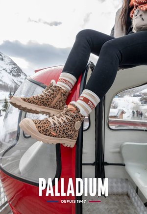 Tansfarbene Leopardenmuster-Ankle Boots mit beigen Schnürsenkeln und Gummisohlen, kombiniert mit gestreiften Socken, vor einem roten Gondel-Hintergrund.