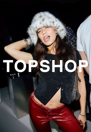 Junge Frau mit weißem Pelzhut, schwarzem bauchfreiem Top und roter Lederhose posiert selbstbewusst auf einer Party mit dem Schriftzug "TOPSHOP".