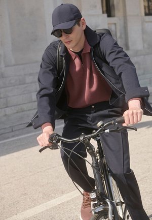 Mann trägt dunkle Jacke, burgunderfarbenes Hemd, Mütze und Sonnenbrille und fährt ein schwarzes Fahrrad auf einem gepflasterten Weg nahe steinerner Stufen.