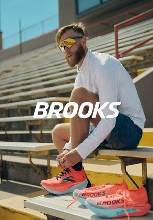 Zapatillas de correr Brooks de un brillante color rosa con detalles en negro, diseño ligero y parte superior de malla texturizada. Camiseta de manga larga blanca y pantalones cortos azul marino llevados por un hombre.