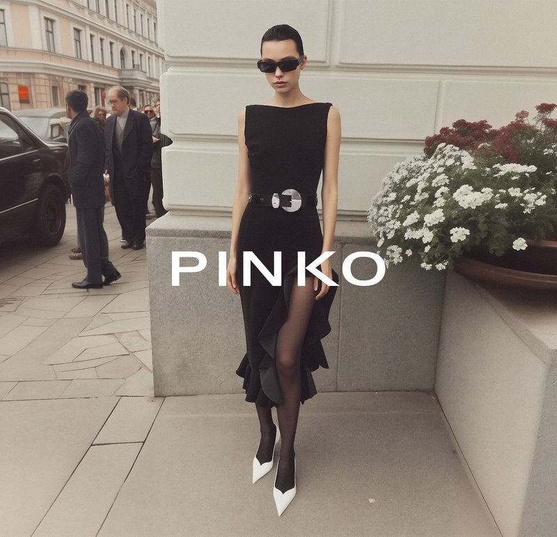 Vrouw in een zwarte mouwloze jurk met ruches, zwarte kousen, witte puntige schoenen en een zonnebril, staand bij een stenen muur met bloemen in de buurt.