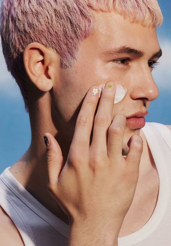 Une personne ayant les cheveux roses applique de la crème sur sa joue, portant un débardeur blanc et montrant des ongles décorés avec des motifs de fleurs et de smileys.