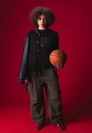 Veste de baseball noire avec des manches en cuir, texte graphique, pantalon cargo vert, baskets rouges, tenant un ballon de basket, fond rouge.