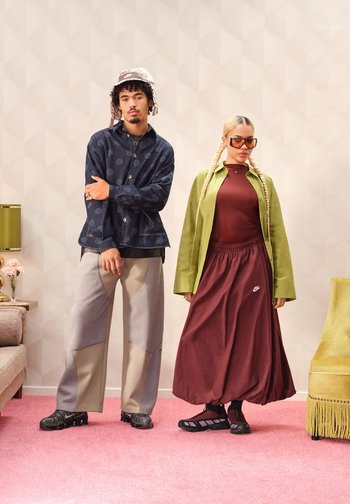 Una persona con una camicia scura a motivi e pantaloni larghi grigio chiaro; un'altra con una giacca verde, un top color marsala e una gonna lunga color marsala, con occhiali da sole.