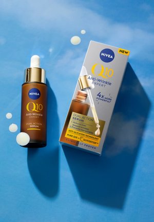 Bottiglia di siero Nivea Q10 Anti-Wrinkle Expert Dual Action accanto alla sua confezione su una superficie blu, con gocce di siero vicine.