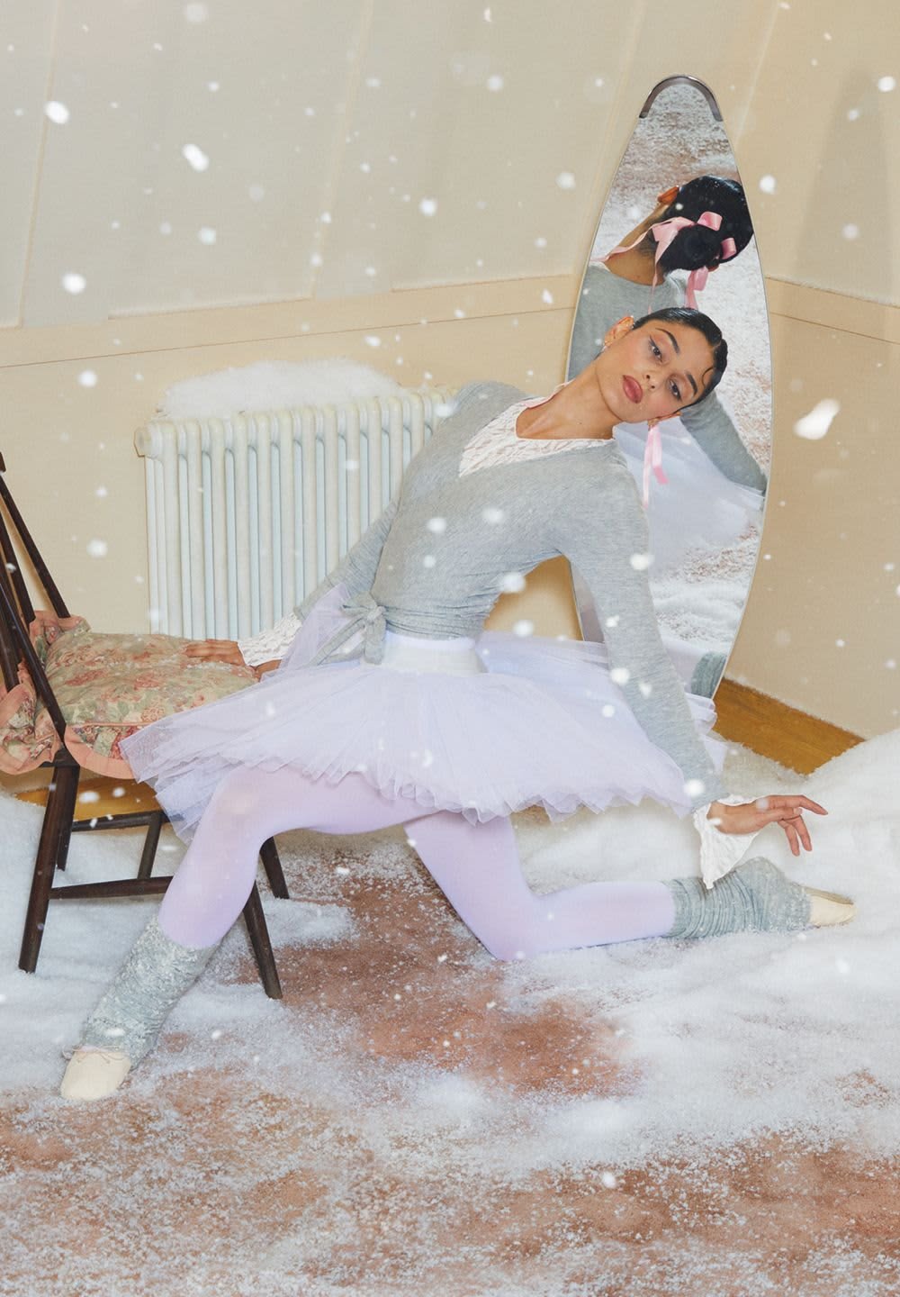 Ballet dancer in a grey wrap top, white tulle skirt, and lavender tights. Grey leg warmers and pointe shoes, posed in a snow-covered room.