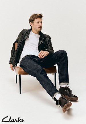 Braune Lederanzugboots mit strukturierter Sohle, dunkle Jeans, ein weißes T-Shirt und eine schwarze Lederjacke auf einer sitzenden Figur in einem Stuhl.