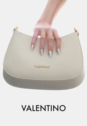 Main avec des ongles vert menthe métallisé tenant un sac à main Valentino beige avec logo doré sur fond blanc.