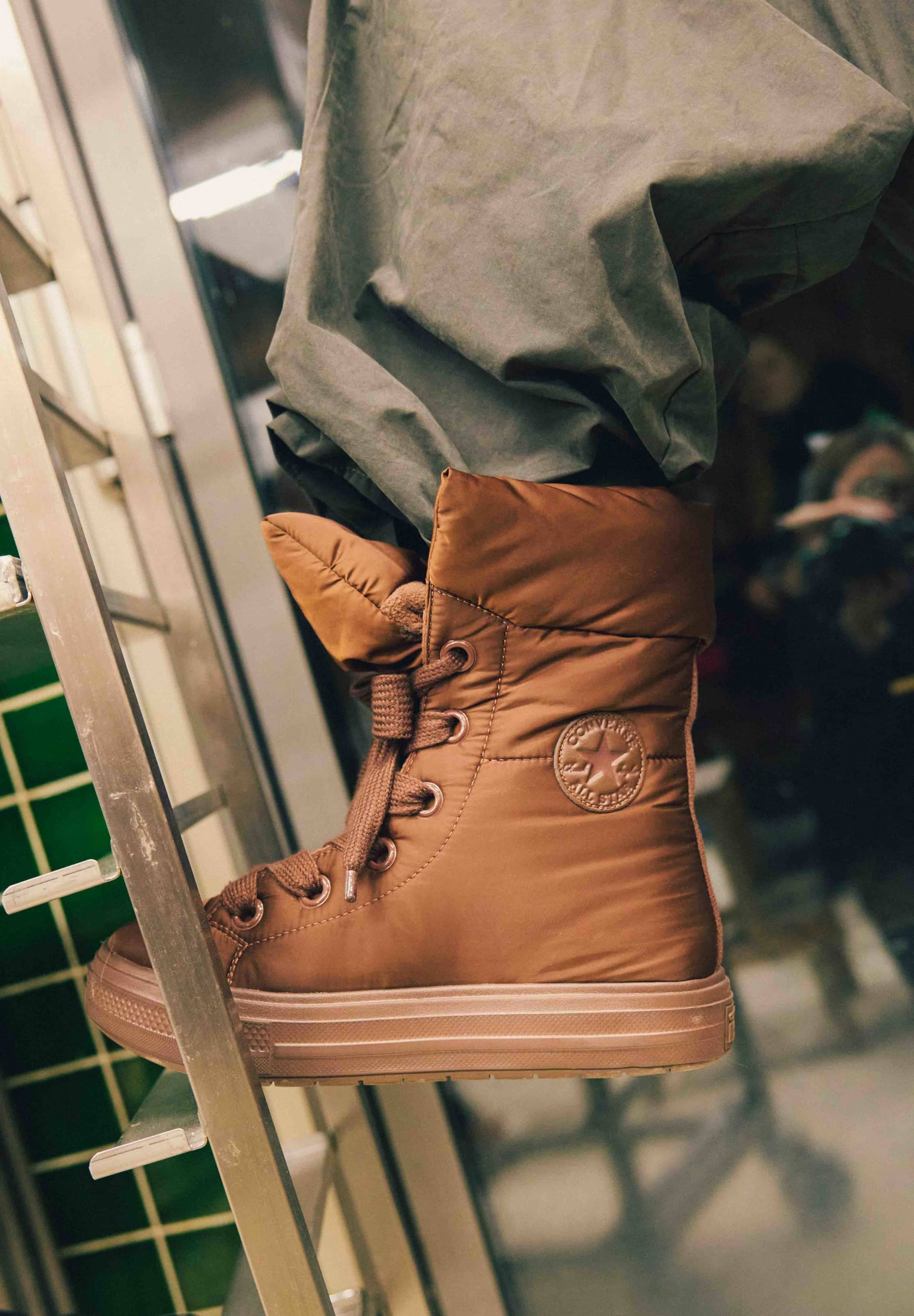 Zapatilla de corte alto acolchada de color marrón con superficie texturizada, puntera redonda de goma y diseño con cordones, con un logotipo circular en el costado.